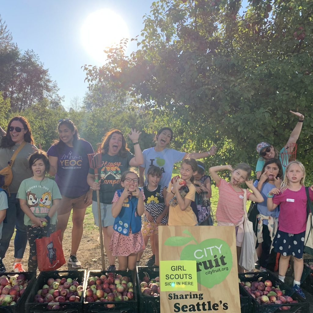 The image shows a group of schoolchildren and their teachers posing in front of an orchard.