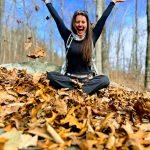 Meg is wearing black pants and shirt. She has long brown hair and is sitting cross legged on a forest floor. She is throwing fallen leaves above her head.