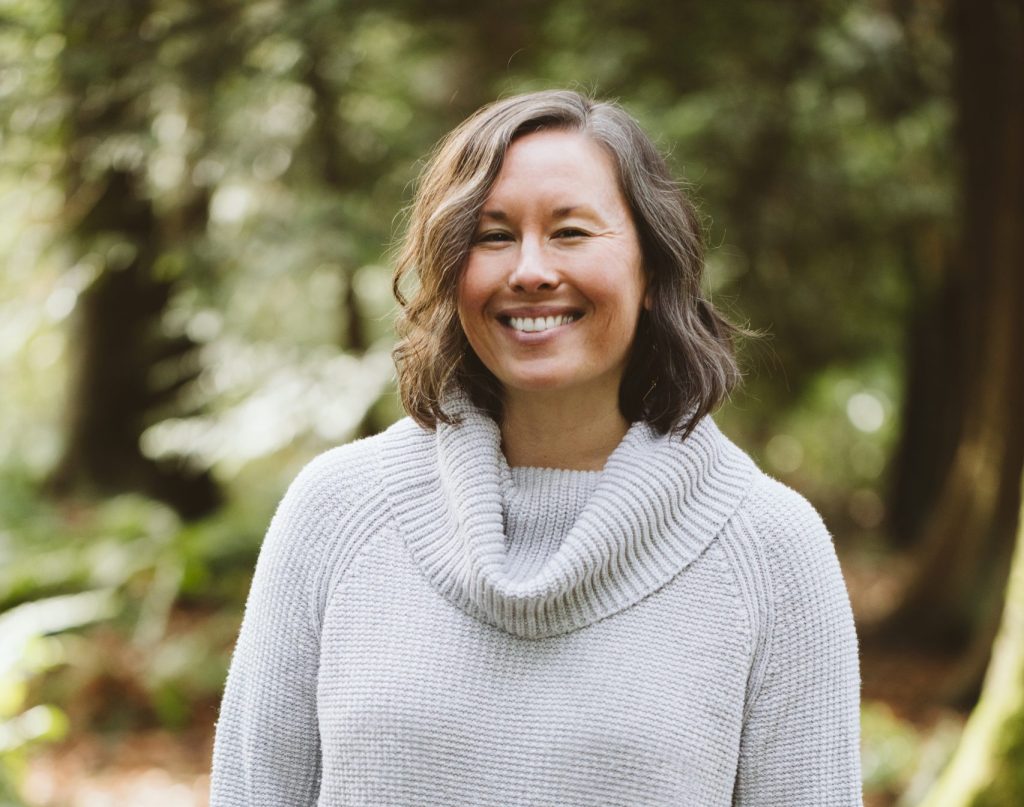 Pictured is City Fruit's Executive Director, Emily Tomita! She is smiling and wearing a grey sweater. She is standing against a green background of trees.