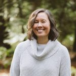 Pictured is City Fruit's Executive Director, Emily Tomita! She is smiling and wearing a grey sweater. She is standing against a green background of trees.