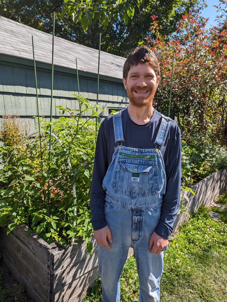 The image shows our tree care specialist Jeff standing in a garden. He has brown curly hair and a beard and is wearing a black cap, a black shirt, and denim overalls.