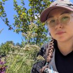 The image depicts Fruit Tree Specialist Izzy. She is wearing a pink hat, glasses, and braids. She is smiling next to some flowers.